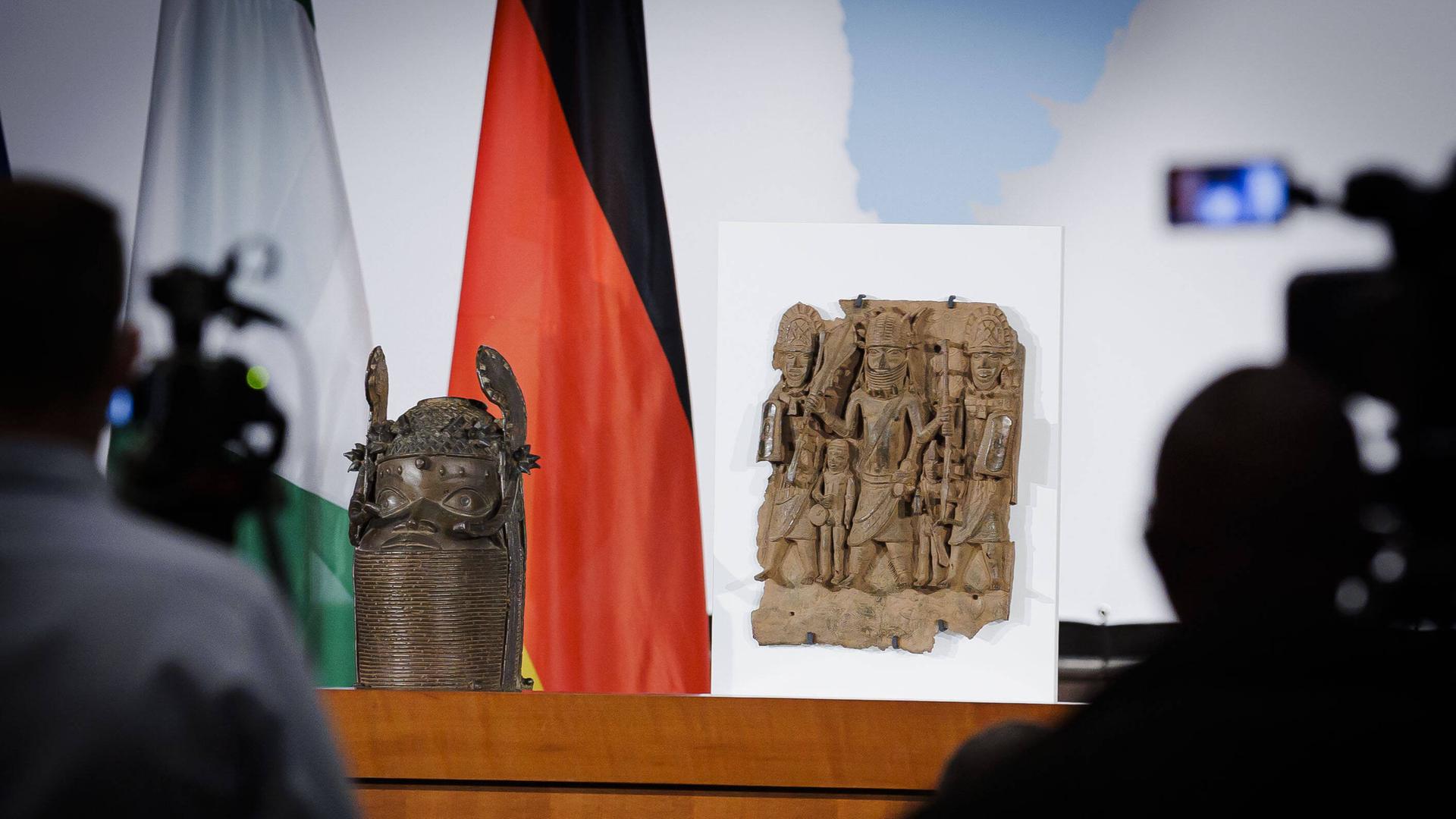 Ein verzierter Kopf aus Bronze und ein Relief mit mehrern Figuren stehen auf einem Tisch, im Hintergrund sind die Fahne Deutschlands und Nigerias. 