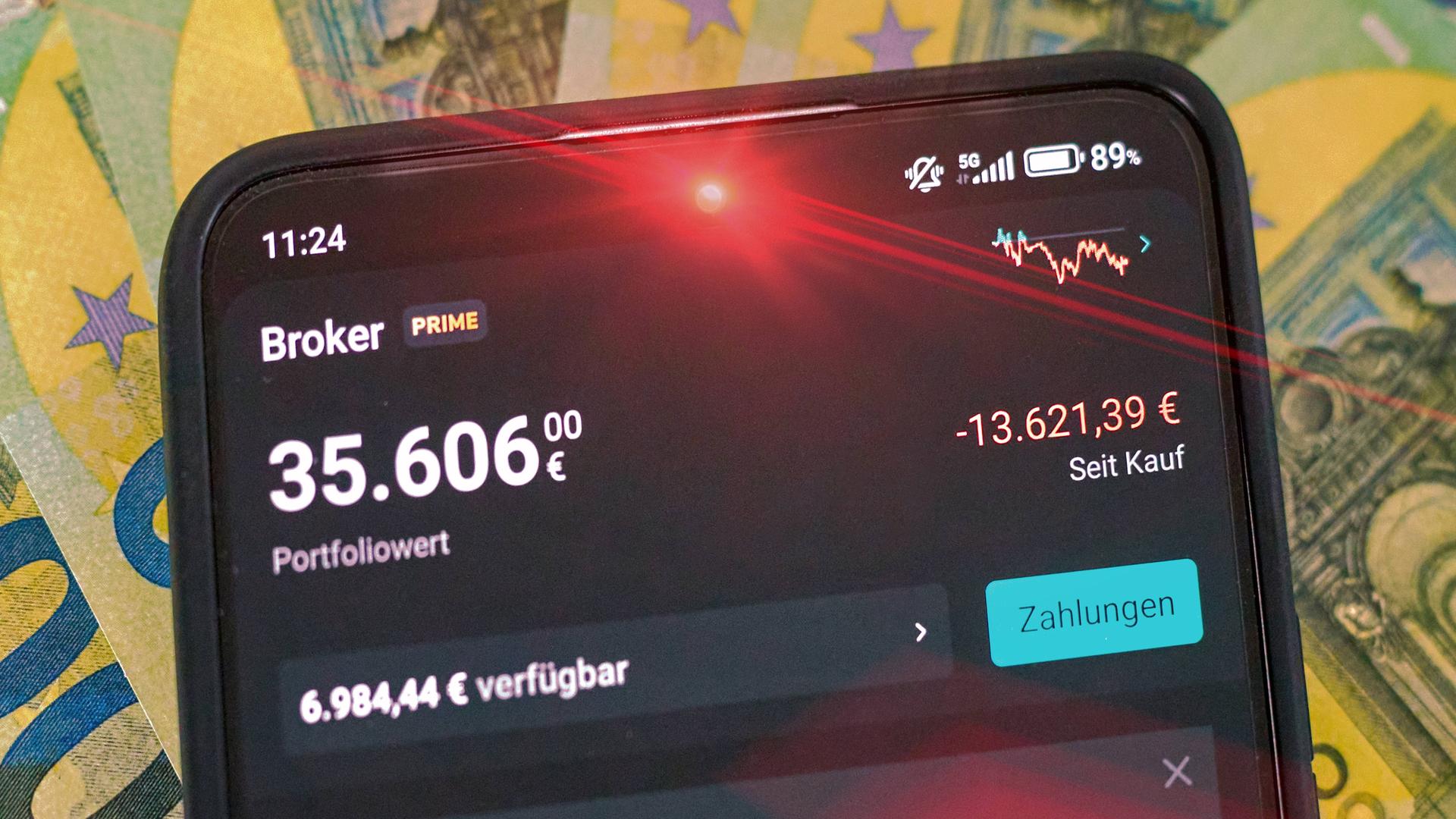Rot leuchtendes Smartphone mit hohem 5-stelligem Geldbetrag, auf dem Display erschein das Portfolio bei Scalable Capital mit dem gesunkenen Portfoliowert. Im Hintergrund liegt Bargeld in 100-Euro-Scheinen.