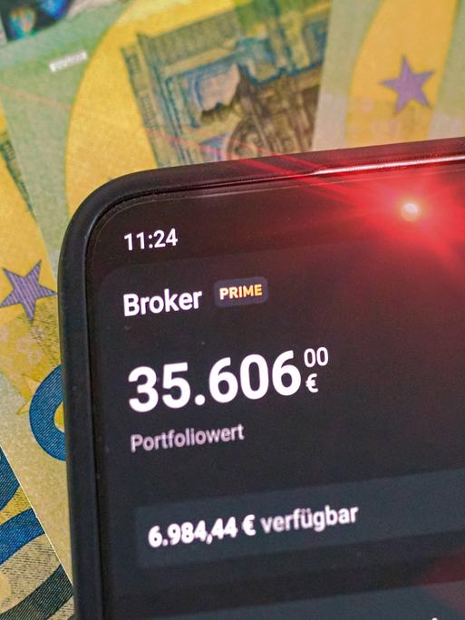 Rot leuchtendes Smartphone mit hohem 5-stelligem Geldbetrag, auf dem Display erschein das Portfolio bei Scalable Capital mit dem gesunkenen Portfoliowert. Im Hintergrund liegt Bargeld in 100-Euro-Scheinen.