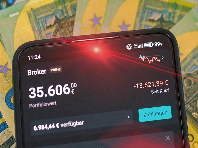 Rot leuchtendes Smartphone mit hohem 5-stelligem Geldbetrag, auf dem Display erschein das Portfolio bei Scalable Capital mit dem gesunkenen Portfoliowert. Im Hintergrund liegt Bargeld in 100-Euro-Scheinen.