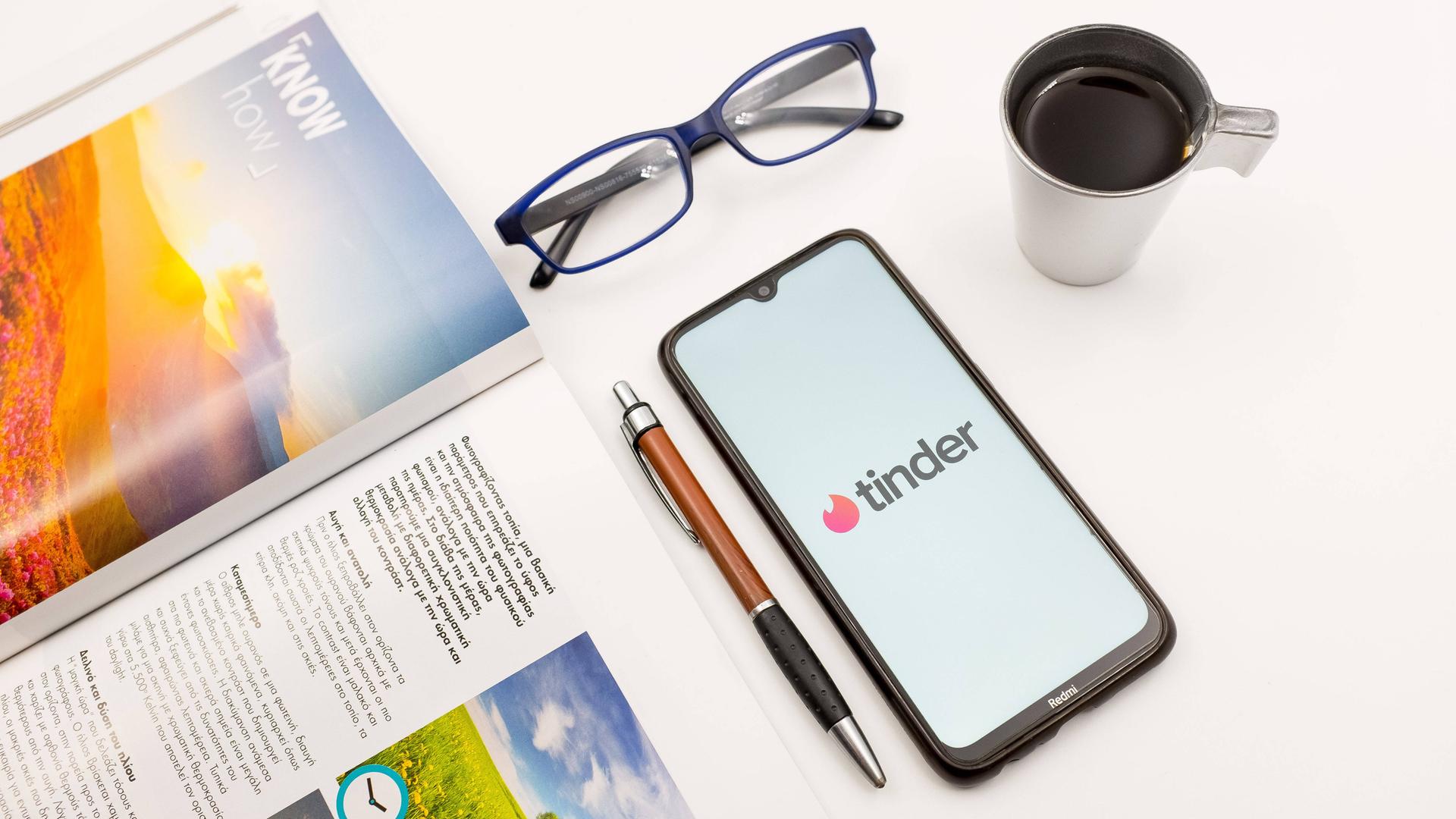 Ein Tisch mit einem Magazin, einem Stift, einer Briller und einem Smartphone auf dem Tinder zu sehen ist.