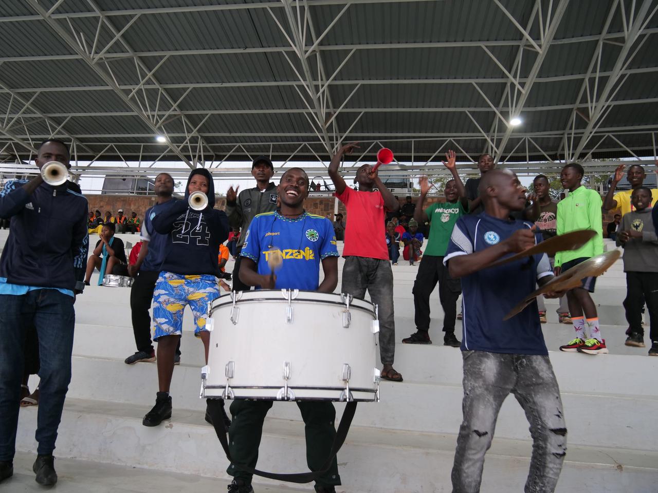 Auf dem Foto sieht man eine Gruppe gut gelaunter ruandischer Fans, die auf der Tribüne stehen und mit Instrumenten wie Tröten und Trommeln die Fußballer anfeuern. 