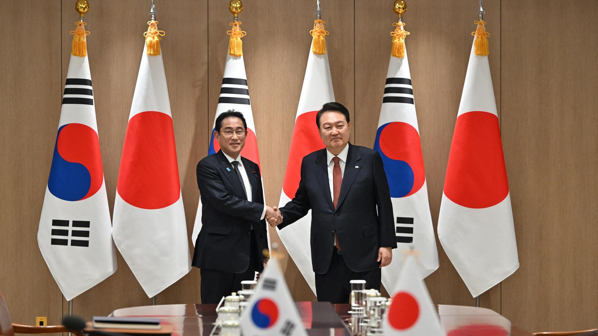 Japans Ministerpräsident Kishida (l.) trifft den südkoreanischen Präsidenten Yoon in Seoul. Beide Politiker tragen einen schwarzen Anzug und schütteln sich die Hände während sie in einem Konferenzraum stehen; im Hintergrund sind die beiden Landesflaggen zu sehen.