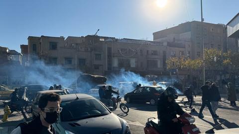 Das Bild zeigt eine Straßenszene in Teheran. Menchen in Autos und auf Motorrädern protestieren. Im Hintergrund sieht man Qualm.