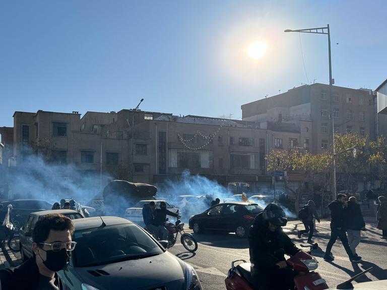 Das Bild zeigt eine Straßenszene in Teheran. Menschen in Autos und auf Motorrädern protestieren. Im Hintergrund sieht man Qualm.