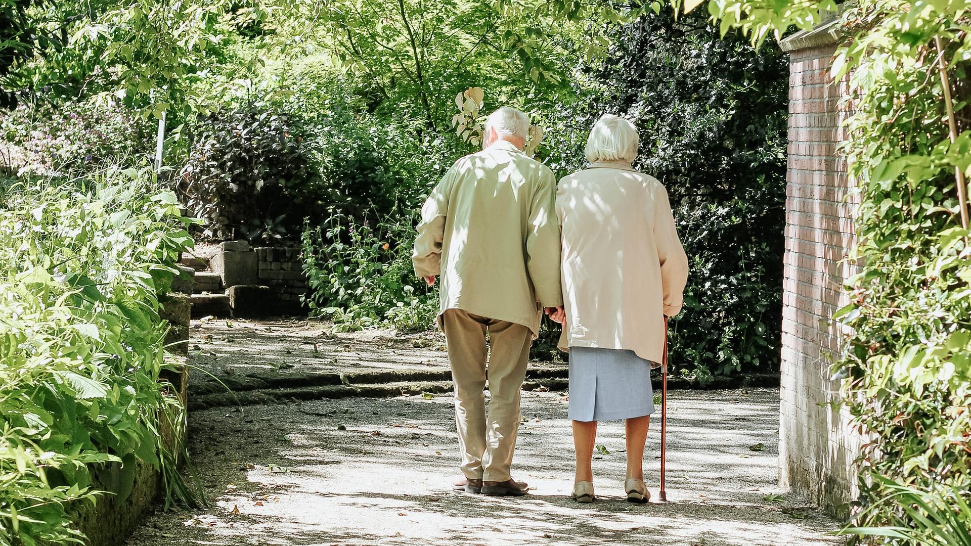 Zwei ältere Personen laufen Hand in Hand über einen Weg, umgeben von Grün.