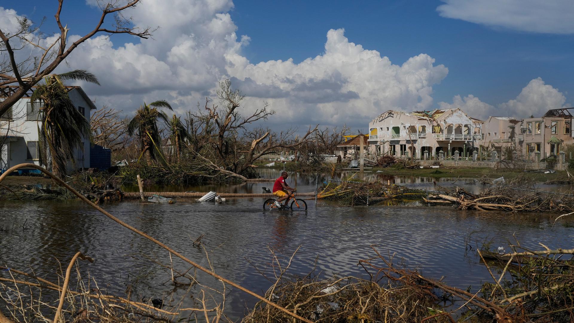 Ein Mann radelt durch eine überflutete Straße, im Hintergrund zerstörte Häuser und blauer Himmel mit weißen Wolken Ein Mann radelt durch eine überflutete Straße, im Hintergrund zerstörte Häuser und blauer Himmel mit weißen Wolken