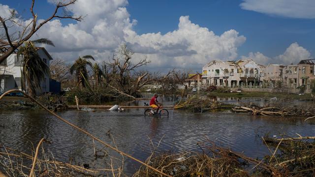 Ein Mann radelt durch eine überflutete Straße, im Hintergrund zerstörte Häuser und blauer Himmel mit weißen Wolken