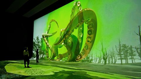 Eine Projektion in leuchtend grüner Farbe in einem sonst verdunkelten großen Raum zeigt Schlangen. Zwei Betrachterinnen schauen auf die Leinwand. Installationsansicht NXT Museum Amsterdam
