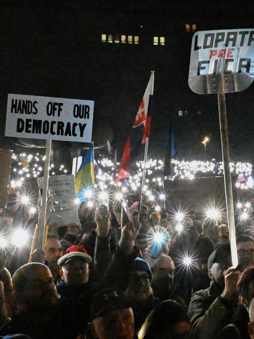 Viele Menschen halten bei einer Demonstration in der Dunkelheit in Bratislava ihre Handylichter nach oben und tragen Plakate, auf einem steht: "Hands off our democracy."