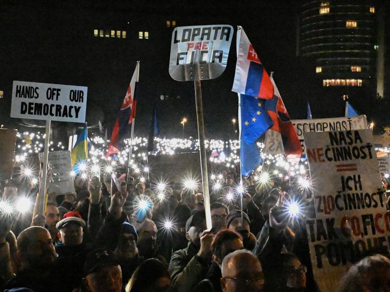 Viele Menschen halten bei einer Demonstration in der Dunkelheit in Bratislava ihre Handylichter nach oben und tragen Plakate, auf einem steht: "Hands off our democracy."