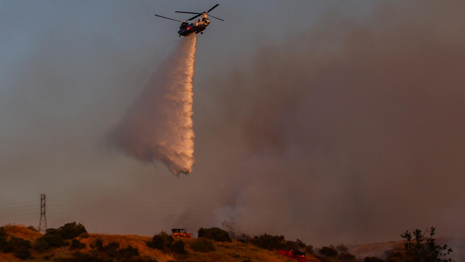 Ein Helikopter schüttet Wasser über einem Waldbrand im Simi Valley in Kalifornien aus. Im Hintergrund sind Rauchwolken zu sehen, der Himmel ist rötlich.