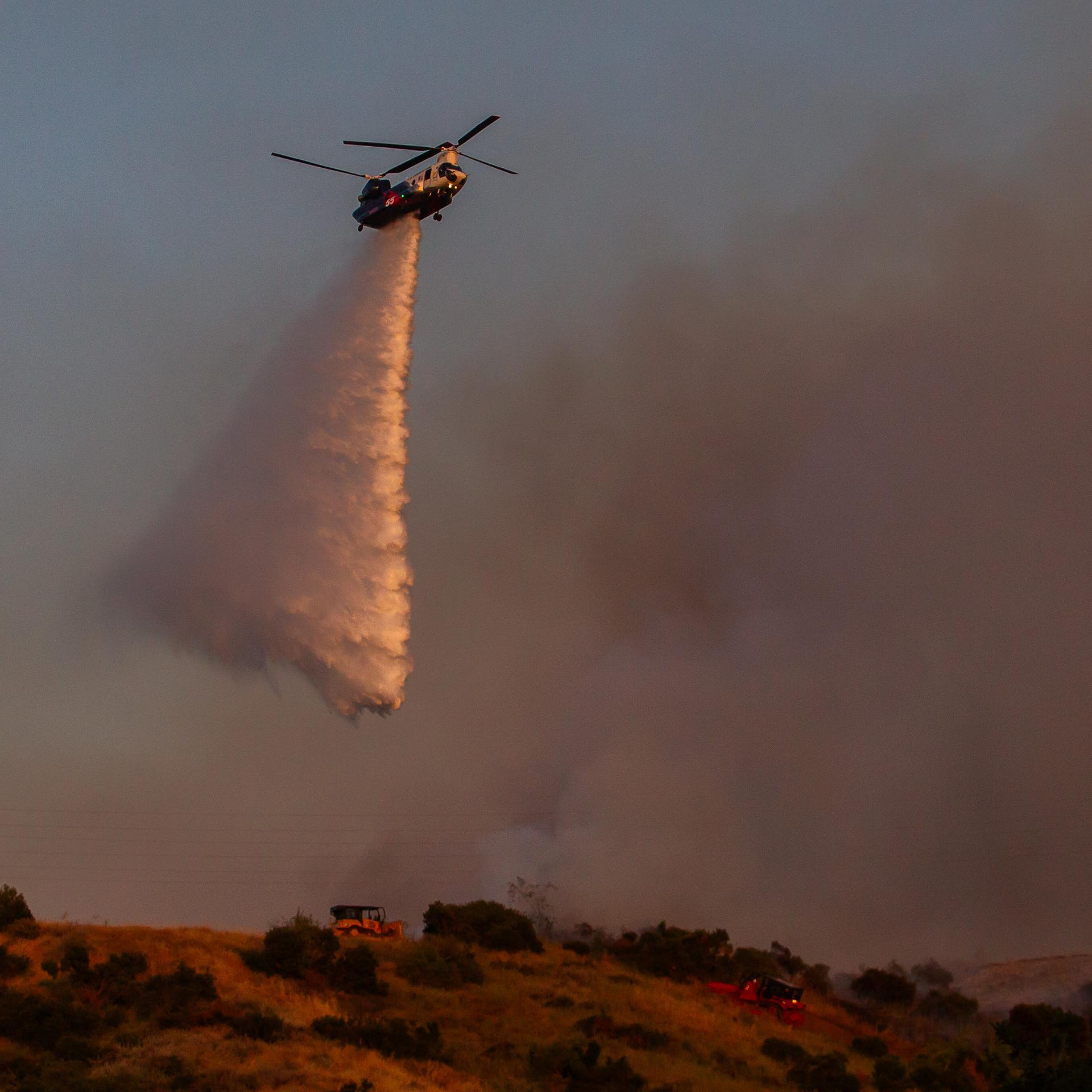 Ein Helikopter schüttet Wasser über einem Waldbrand im Simi Valley in Kalifornien aus. Im Hintergrund sind Rauchwolken zu sehen, der Himmel ist rötlich.