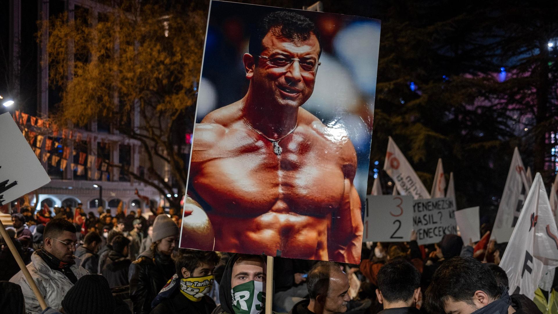 Ein junger vermummter Demonstrant hält ein Schild in de Höhe. Auf dem Schild ist der Ekrem Imamoglu als Bodybuilder zu sehen.