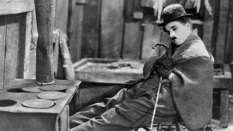 Film Still aus "The Gold Rush" (1925): Charlie Chaplin mit ikonographischem Oberlippenbart und Bowler, sitzt mit geschlossenen Augen auf einem Stuhl, eine Decke um die Schultern gelegt. Film Still aus "The Gold Rush" (1925): Charlie Chaplin mit ikonographischem Oberlippenbart und Bowler, sitzt mit geschlossenen Augen auf einem Stuhl, eine Decke um die Schultern gelegt.