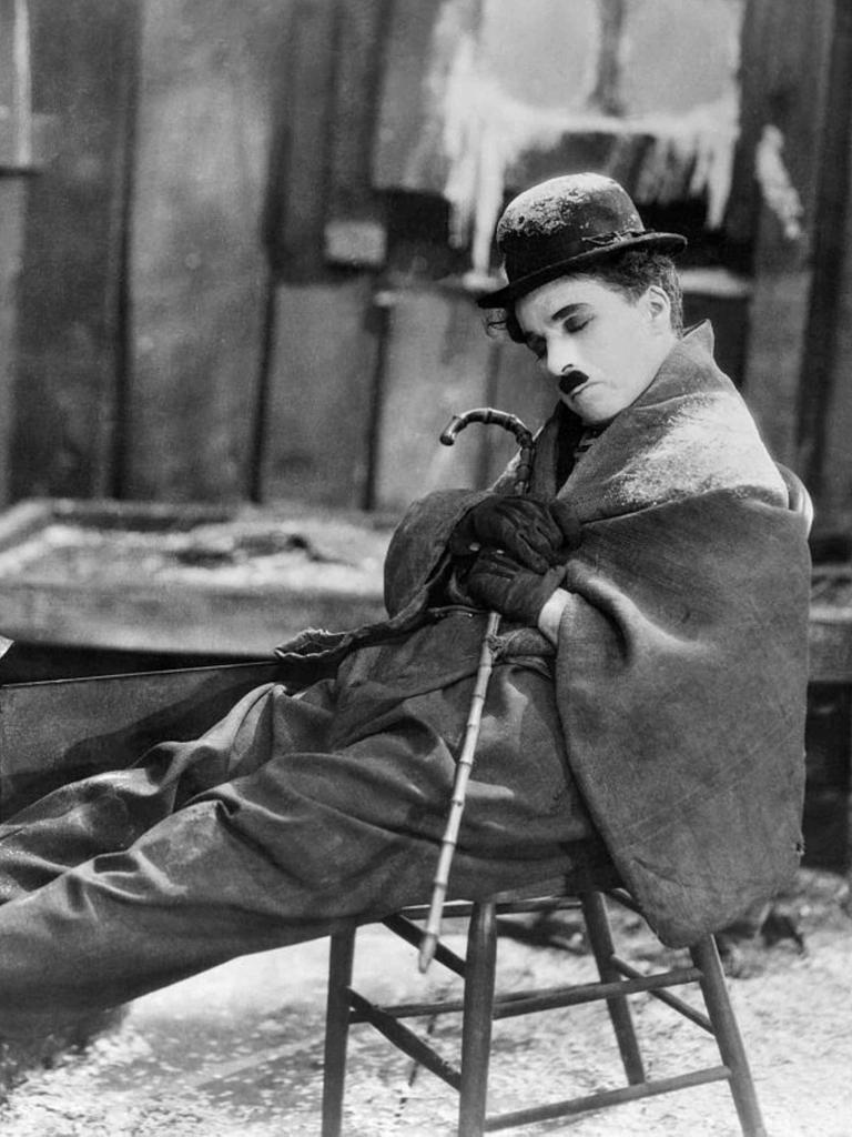 Film Still aus "The Gold Rush" (1925): Charlie Chaplin mit ikonographischem Oberlippenbart und Bowler, sitzt mit geschlossenen Augen auf einem Stuhl, eine Decke um die Schultern gelegt.
