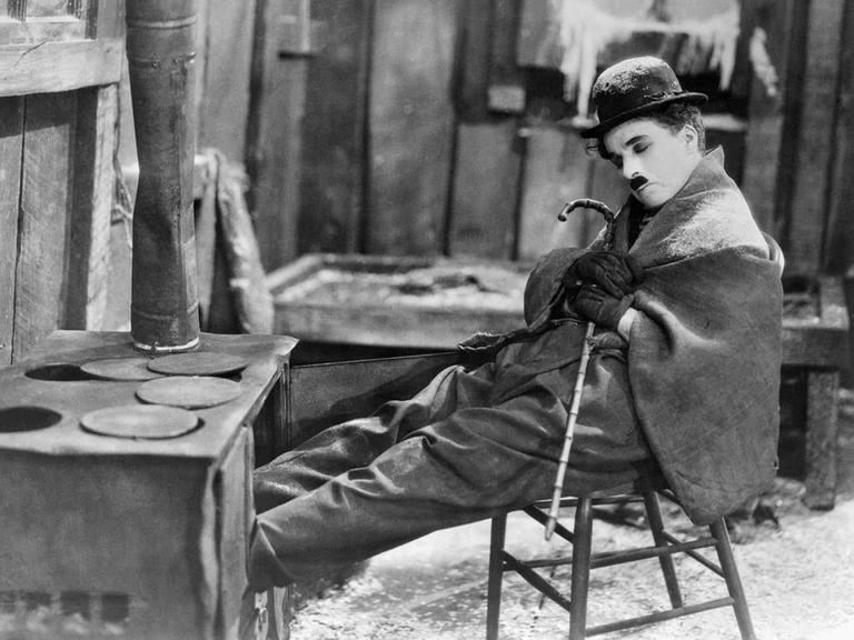 Film Still aus "The Gold Rush" (1925): Charlie Chaplin mit ikonographischem Oberlippenbart und Bowler, sitzt mit geschlossenen Augen auf einem Stuhl, eine Decke um die Schultern gelegt. Film Still aus "The Gold Rush" (1925): Charlie Chaplin mit ikonographischem Oberlippenbart und Bowler, sitzt mit geschlossenen Augen auf einem Stuhl, eine Decke um die Schultern gelegt.
