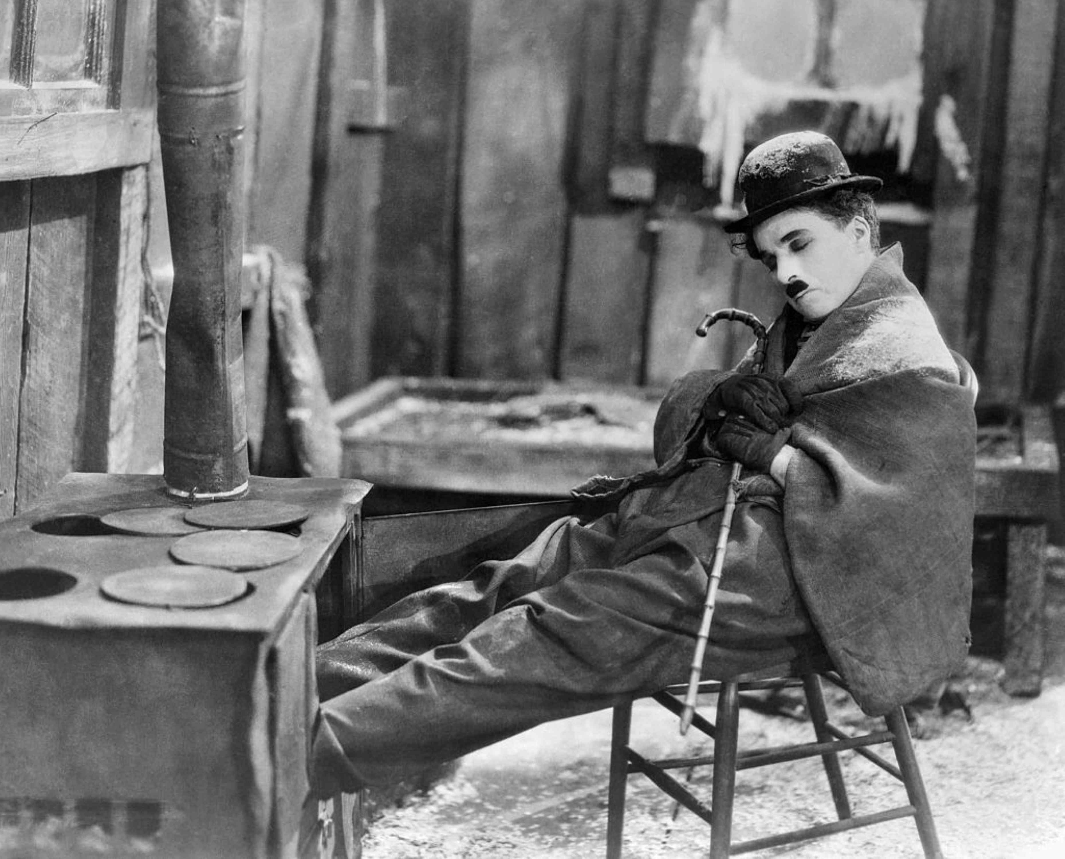Film Still aus "The Gold Rush" (1925): Charlie Chaplin mit ikonographischem Oberlippenbart und Bowler, sitzt mit geschlossenen Augen auf einem Stuhl, eine Decke um die Schultern gelegt.