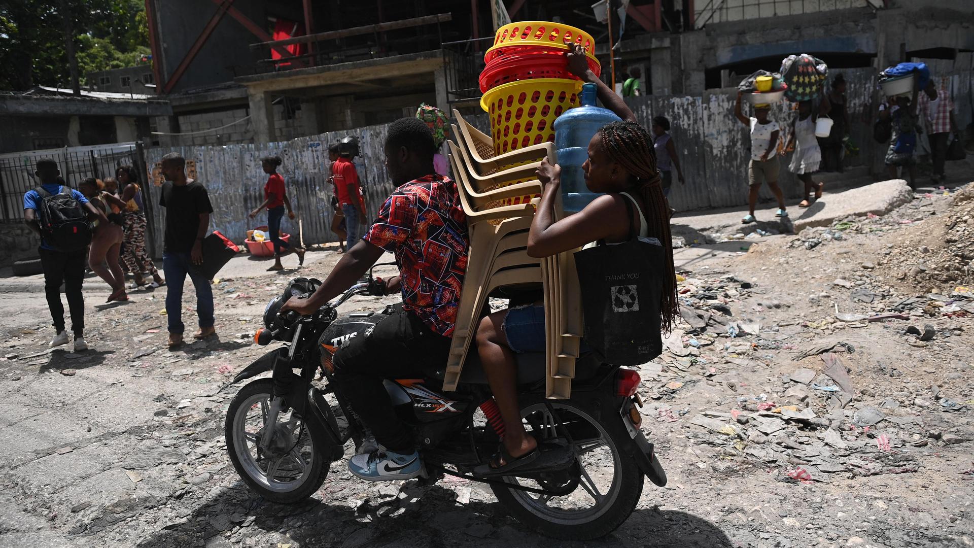 Ein Mann und eine Frau transportieren auf einem Motorrad Stühle, Körbe und eine Gasflasche. Im Hintergrund sind weitere Menschen mit Gepäck zu sehen.