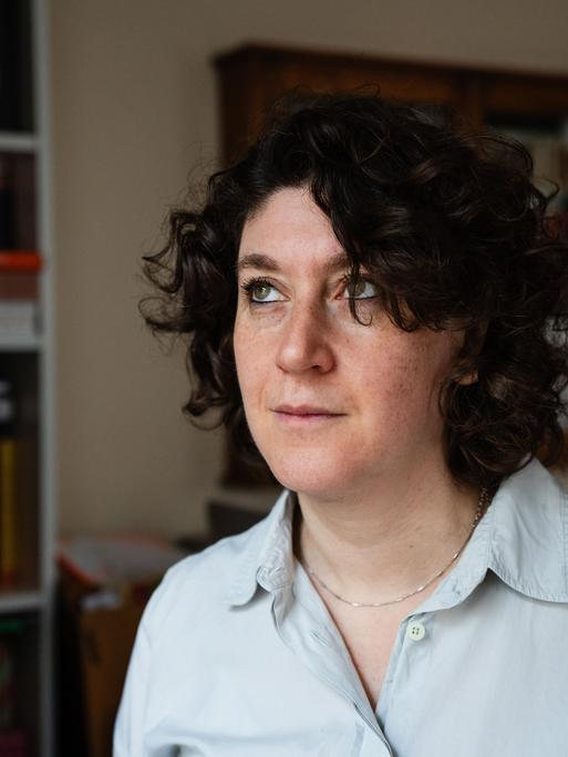 Eine Frau mit dunklen lockigen Haaren trägt ein weißes Hemd. Sie steht in einem Raum und blickt nach oben. Es ist die Autorin Lena Gorelik.