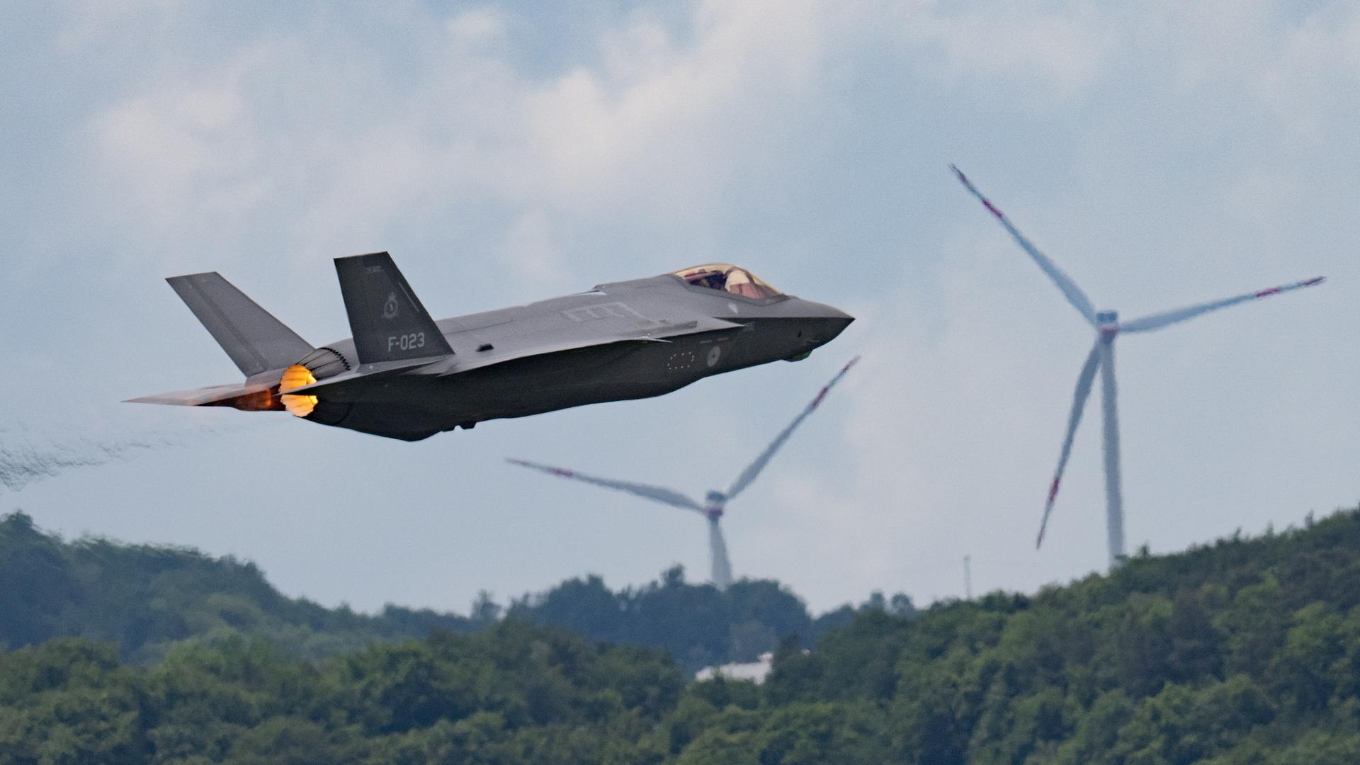 Ein Kampfjet vom Typ F-35 startet über einem Wald und Windrädern von der US-Airbase Ramstein. 