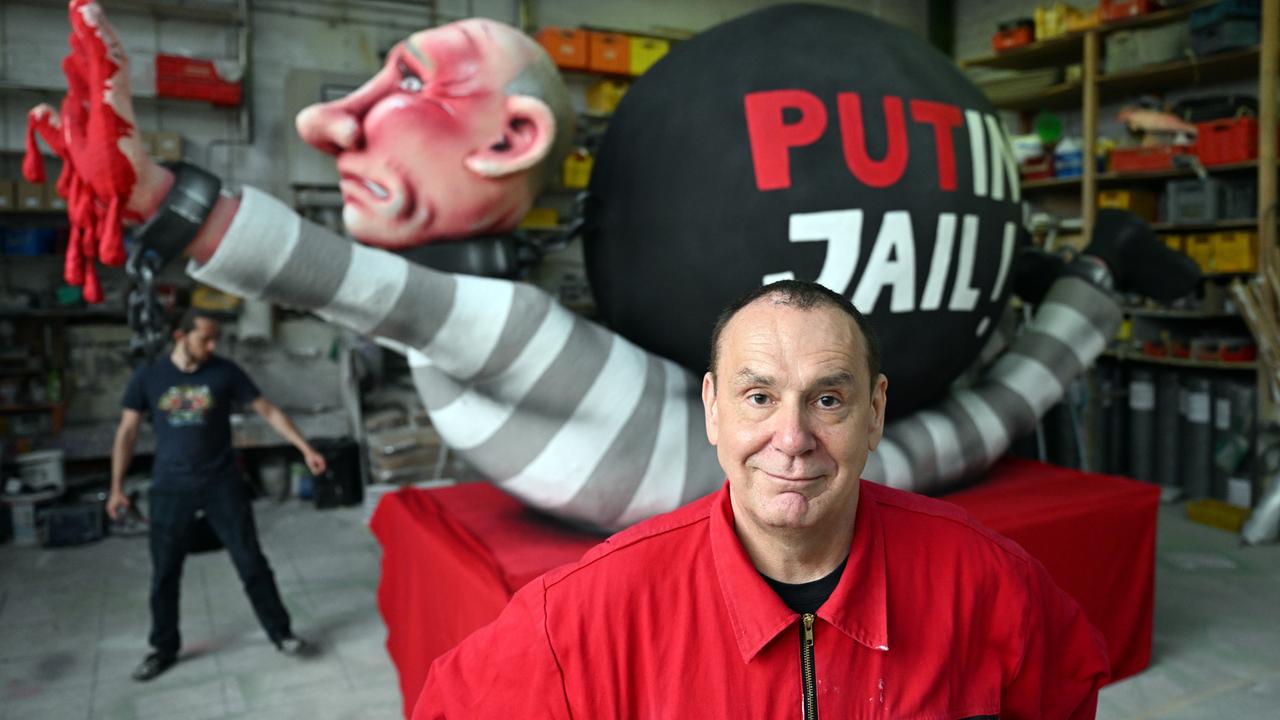 Jacques Tilly trägt einen roten Anzug und steht vor einer Figur, die den russischen Präsidenten Putin mit roten Händen zeigt. Auf der Figur steht "PutIn Jail".