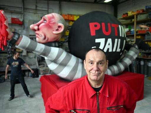 Jacques Tilly trägt einen roten Anzug und steht vor einer Figur, die den russischen Präsidenten Putin mit roten Händen zeigt. Auf der Figur steht "PutIn Jail".