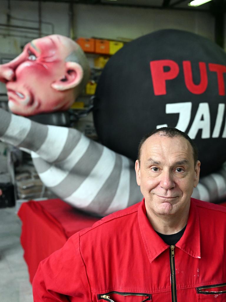 Jacques Tilly trägt einen roten Anzug und steht vor einer Figur, die den russischen Präsidenten Putin mit roten Händen zeigt. Auf der Figur steht "PutIn Jail".