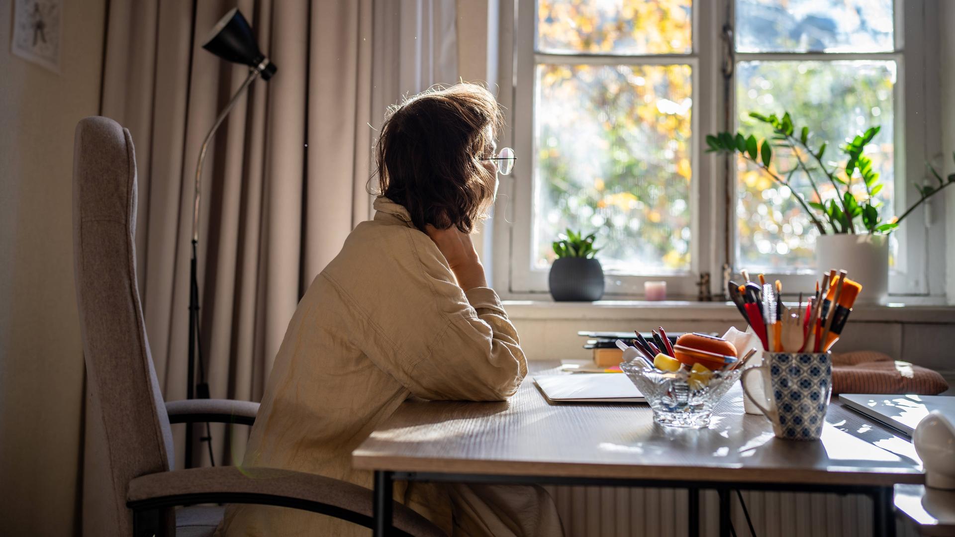Foto einer jungen Frau, die an einem Schreibtisch sitzt und nachdenklich aus dem Fenster schaut. Foto einer jungen Frau, die an einem Schreibtisch sitzt und nachdenklich aus dem Fenster schaut.