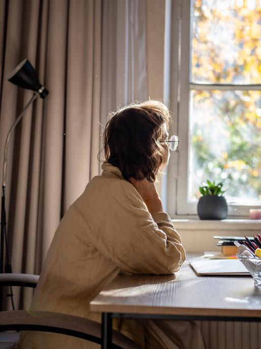 Foto einer jungen Frau, die an einem Schreibtisch sitzt und nachdenklich aus dem Fenster schaut. 