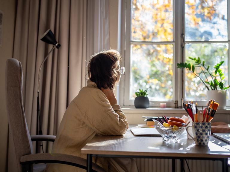 Foto einer jungen Frau, die an einem Schreibtisch sitzt und nachdenklich aus dem Fenster schaut. 