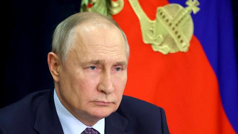 Russlands Präsident Putin sitzt vor einer blau-roten Fahne mit goldenem aufgesticktem Reichsapfel.