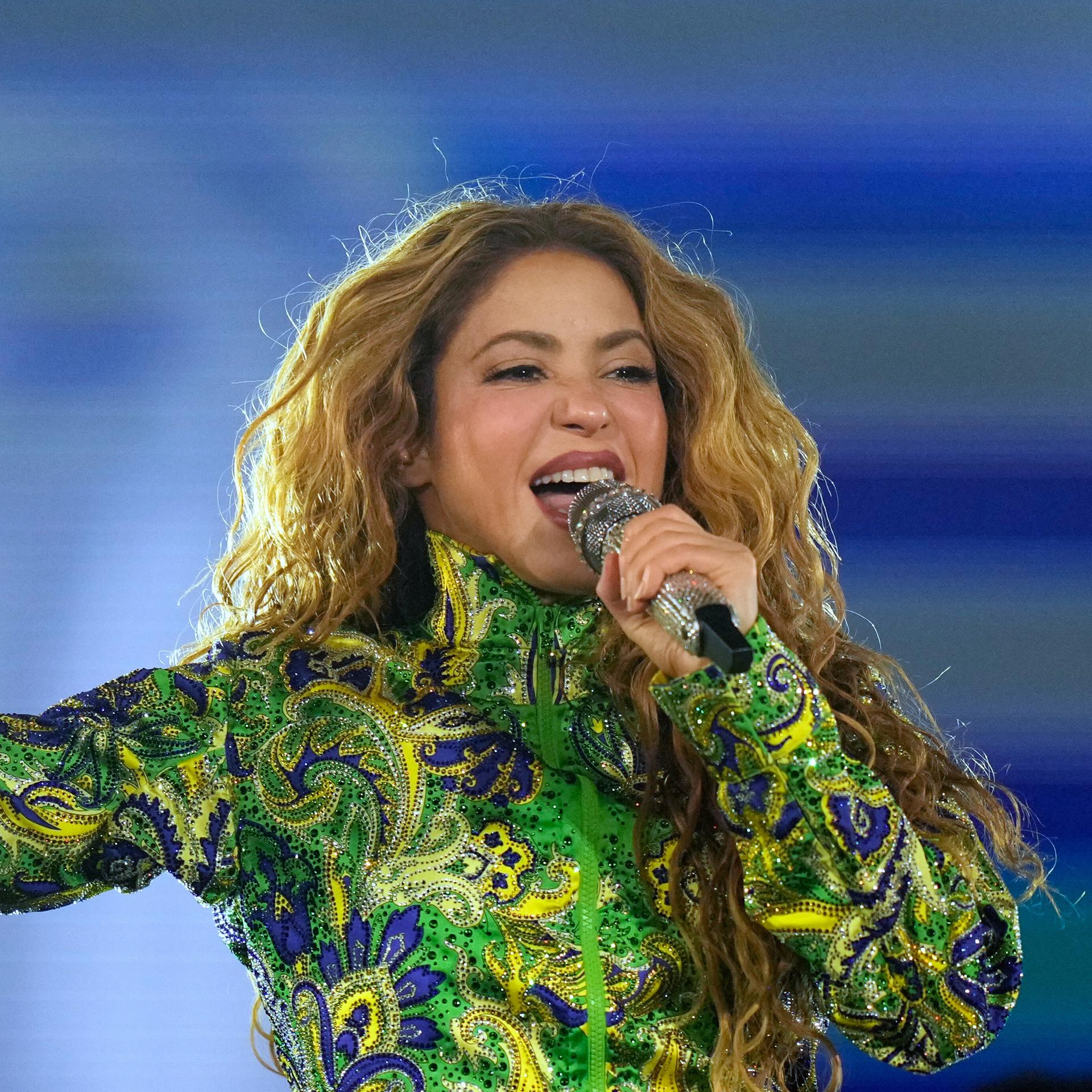 Shakira singt mit einem Mikro vor dem Mund. Sie hat ein langärmeliges Obertteil in grün, blau und gelb an. Die rechte Hand hält sie nach oben, Zeigefinger und kleiner Finger sind nach oben gereckt.