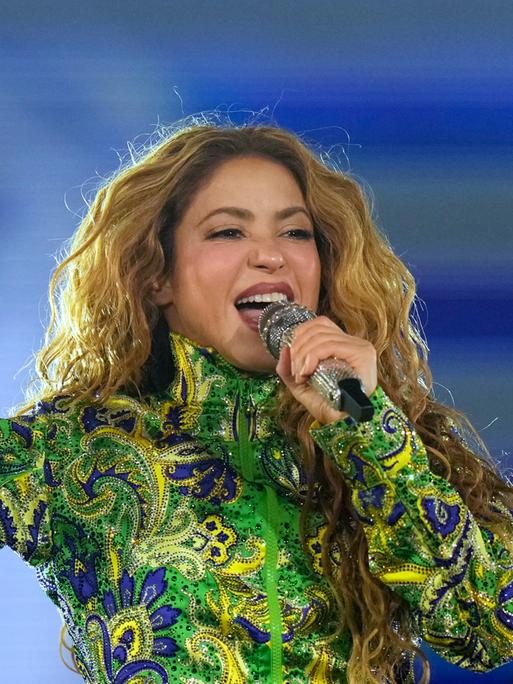 Shakira singt mit einem Mikro vor dem Mund. Sie hat ein langärmeliges Obertteil in grün, blau und gelb an. Die rechte Hand hält sie nach oben, Zeigefinger und kleiner Finger sind nach oben gereckt.
