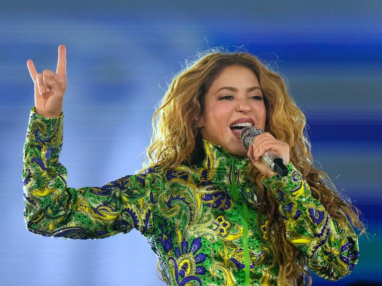 Shakira singt mit einem Mikro vor dem Mund. Sie hat ein langärmeliges Obertteil in grün, blau und gelb an. Die rechte Hand hält sie nach oben, Zeigefinger und kleiner Finger sind nach oben gereckt.