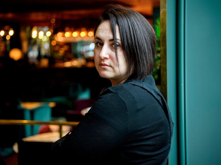 Nino Haratischwili schaut über ihre linke Schulter in die Kamera. Rechts von ihr ist eine grüne Holzverkleidung zu sehen, rechts verschwommene Lichter, vielleicht einer Bar. Eine Strähne ihrer schwarzen, etwa kinnlangen Haare fällt ihr über das Auge.