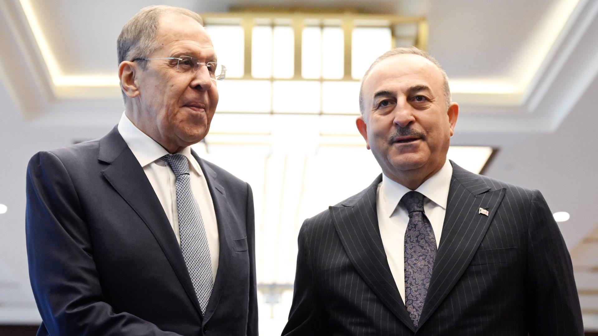 Der russische Außenminister Lawrow steht neben seinem türkischen Kollegen Cavusoglu, sie schütteln sich die Hände und schauen in unterschiedliche Richtungen, beide tragen dunkle Anzüge, weiße Hemden und Krawatten.