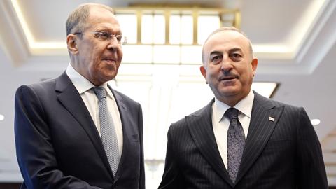 Der russische Außenminister Lawrow steht neben seinem türkischen Kollegen Çavuşoğlu, sie schütteln sich die Hände und schauen in unterschiedliche Richtungen, beide tragen dunkle Anzüge, weiße Hemden und Krawatten.