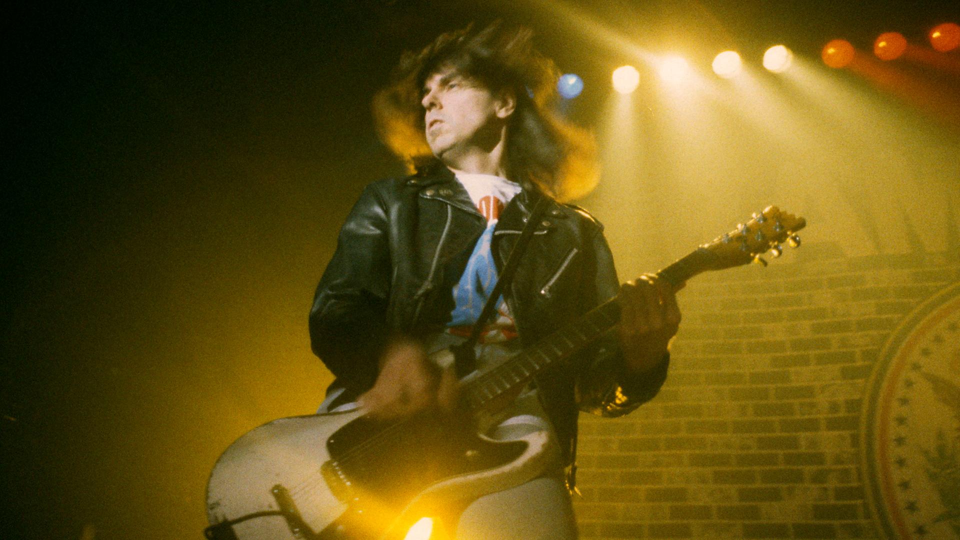 Johnny Ramone von den Ramones live beim SuperBang '93 in der Eilenriedenhalle. Hannover, 12.12.1993. Er spielt dymanisch die Gitarre, seine Haare fliegen im Wind. Die Bühen ist in gelbes Licht geflutet und leicht nebelig. 