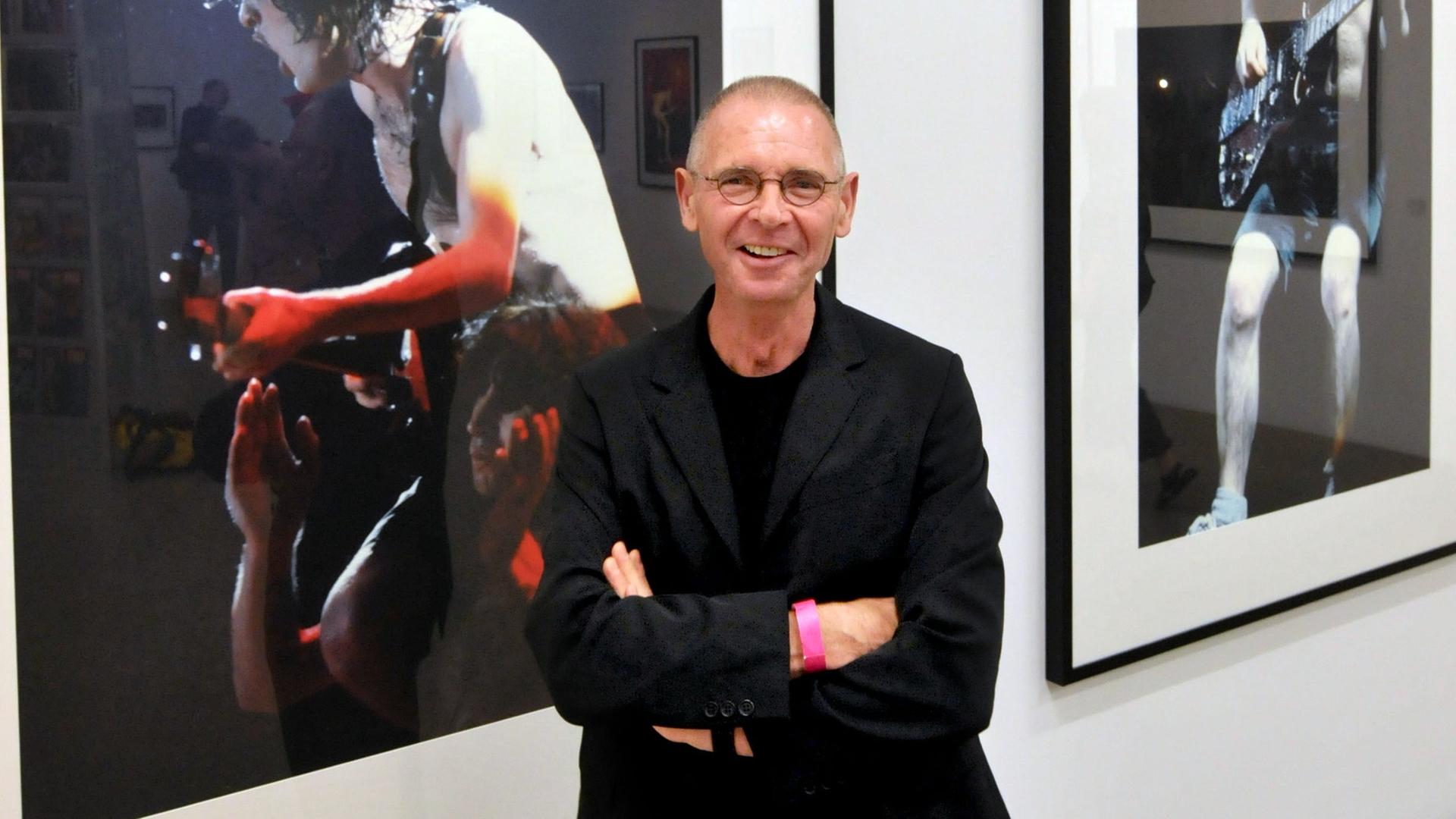 Der Schweizer Fotograf Hannes Schmid trägt eine Brille und lächelt leicht schief. Hinter ihm sind Fotos von einem Rockgitarristen mit freiem Oberkörper zu sehen.