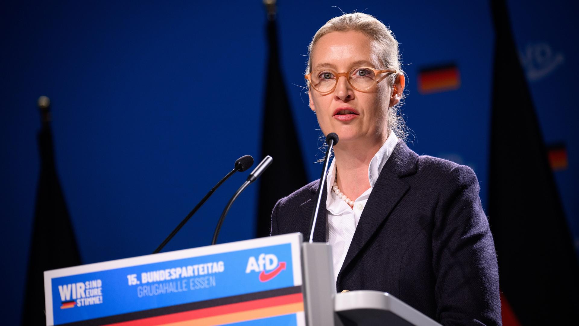 Alice Weidel, eine blonde Frau mit Brille, trägt eine weiße Bluse und einen dunklen Blazer. Sie steht am Rednerpult auf der Bühne vom AfD-Bundesparteitag. Auf dem blauen Pult ist ein AfD-Logo und ein Streifen mit Deutschlandfarben zu sehen.