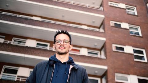 Ein dunkelhaariger junger Mann mit Brille steht vor einem großen Wohnblock.