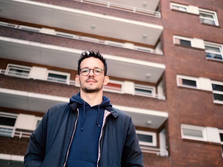 Ein dunkelhaariger junger Mann mit Brille steht vor einem großen Wohnblock.