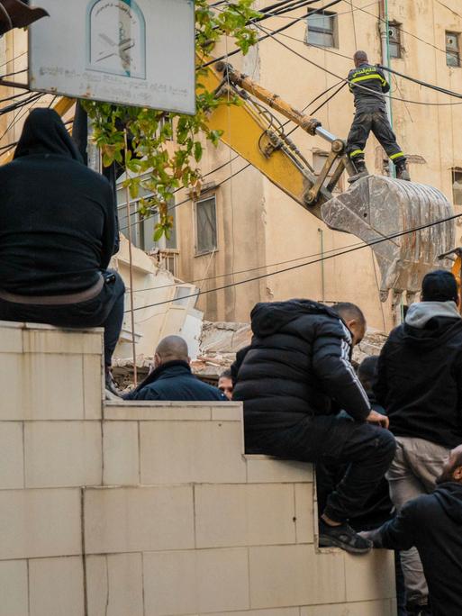 Männer blicken auf ein zerstörtes Wohnhaus, vor dem ein Bagger Schutt wegräumt.