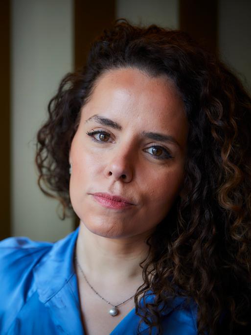 Portrait der Journalistin Alena Jabarine. Sie trägt eine helle blaue Bluse. Ihre langen, lockigen Haare liegen auf ihrer linken Schulter. Sie blickt mit entschlossenem Blick in die Kamera.