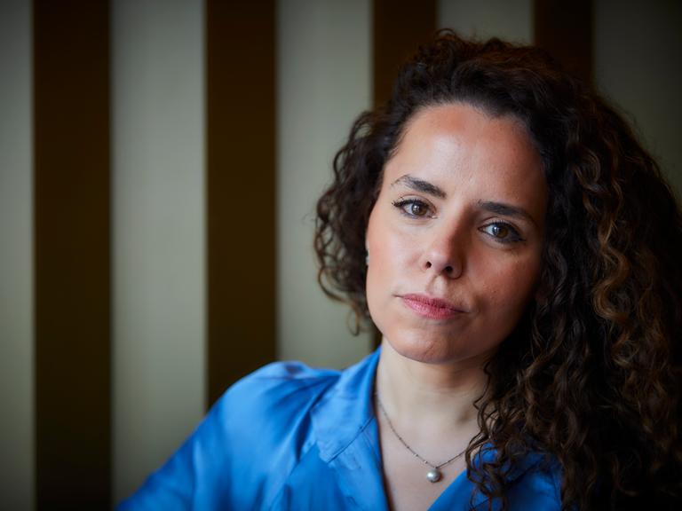 Portrait der Journalistin Alena Jabarine. Sie trägt eine helle blaue Bluse. Ihre langen, lockigen Haare liegen auf ihrer linken Schulter. Sie blickt mit entschlossenem Blick in die Kamera.