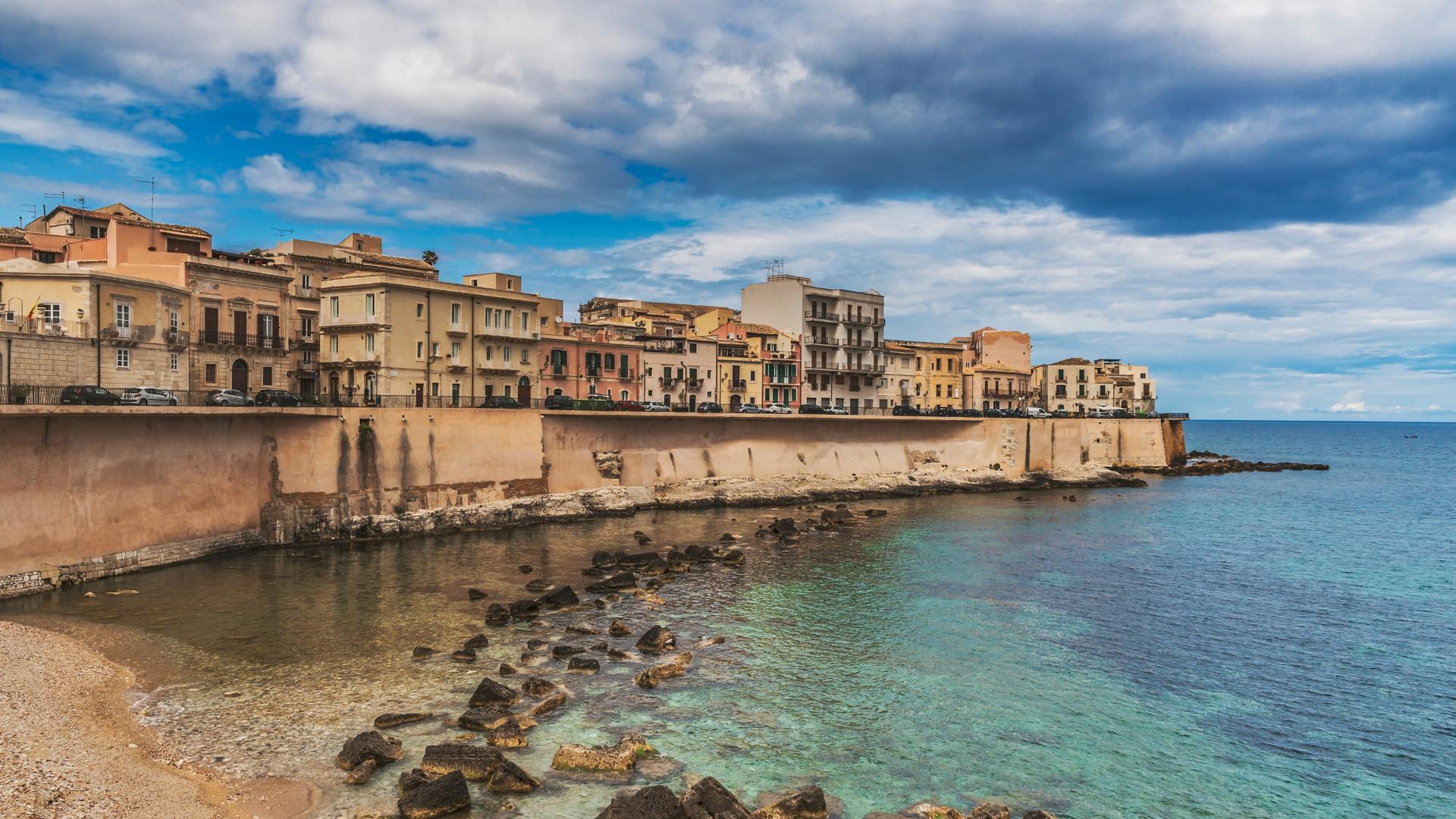 Eine dicht bebaute Landzunge an einem Meer. Eine Mauer trennt das Wasser und die Gebäude. Die Stadt ist Syrakus in Sizilien. 