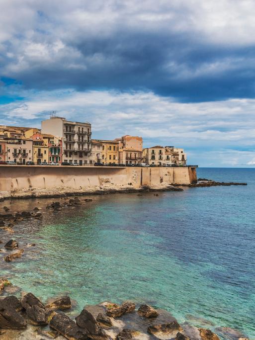 Eine dicht bebaute Landzunge an einem Meer. Eine Mauer trennt das Wasser und die Gebäude. Die Stadt ist Syrakus in Sizilien. 