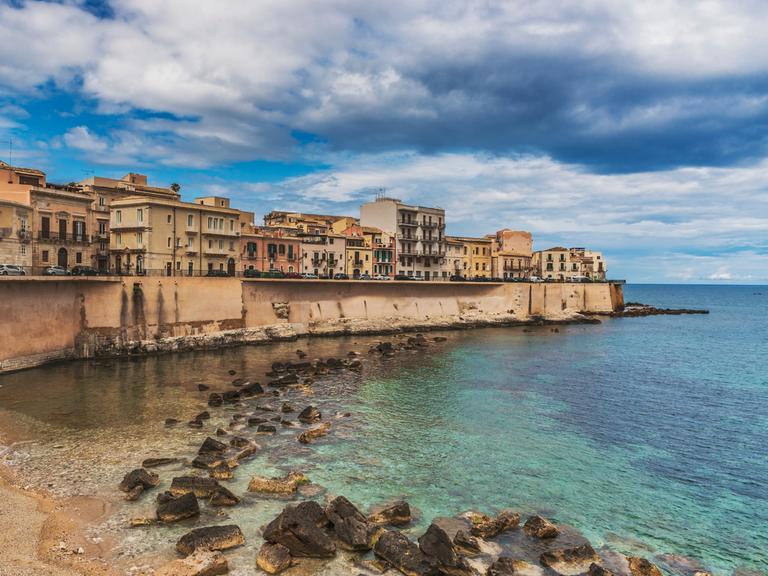 Eine dicht bebaute Landzunge an einem Meer. Eine Mauer trennt das Wasser und die Gebäude. Die Stadt ist Syrakus in Sizilien. 