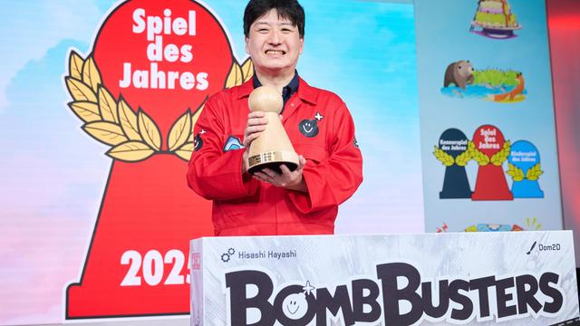 Der Erfinder von dem Spiel "Bomb Busters" heißt Hisashi Hayashi. Er freut sich. Er hält den Preis in den Händen.  